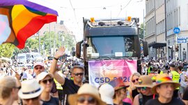 During a Pride parade, people drive through the city in a truck decorated with rainbow flags and celebrate on a sunny street.