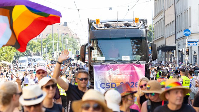 Die Menschen ziehen bei einer Pride-Parade mit einem Lastwagen und Regenbogenfahnen durch die Stadt und feiern auf einer sonnigen Straße.