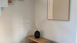 Minimalistischer Eingangsbereich mit Holzbank, runden Kissen, einer schwarzen Vase mit Zweigen und einem großen Wandspiegel.
