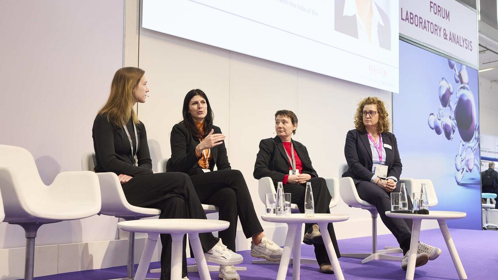 Four women sit on a panel discussion stage, each with a microphone and water glass, in front of a screen.