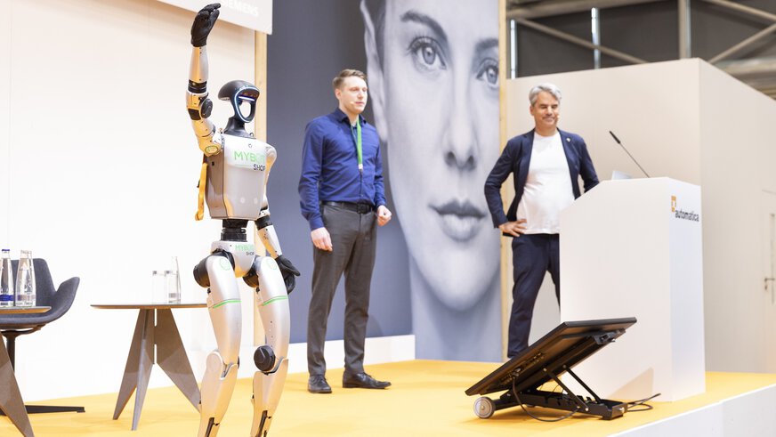 Ein humanoider Roboter mit der Aufschrift „MYBOT SHOP“ steht auf einer Bühne und hebt einen Arm. Zwei Männer stehen daneben, einer am Rednerpult mit dem Logo der automatica.