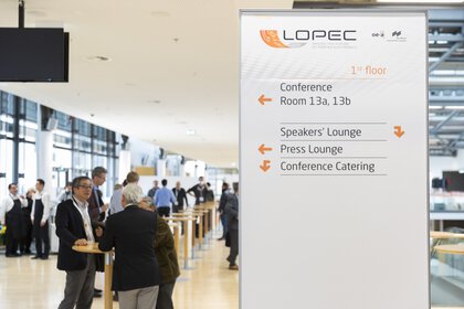 People are socializing next to a conference sign with directions to rooms and lounges.