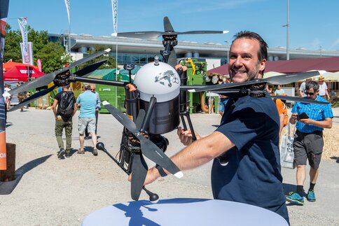 Mann präsentiert eine große Drohne mit mehreren Rotoren auf dem Freigelände einer Fachmesse