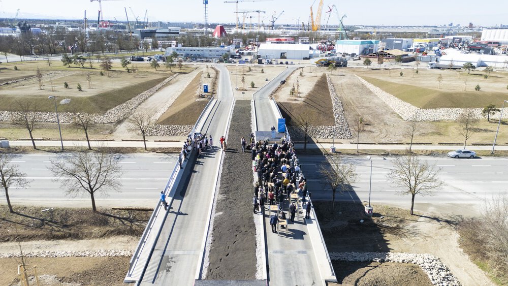 Luftaufnahme einer Menschenmenge, die sich auf einer im Bau befindlichen Brücke inmitten einer Industrielandschaft versammelt hat.