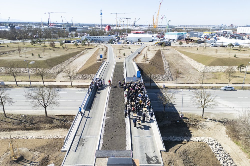 Luftaufnahme einer Menschenmenge, die sich auf einer im Bau befindlichen Brücke inmitten einer Industrielandschaft versammelt hat.