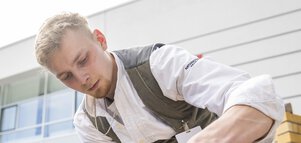 Ein Mann in Kluft baut unter freiem Himmel eine Mauer, während ihm einige Leute dabei zuschauen.