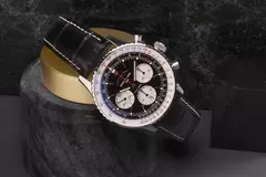 Eine Breitling-Uhr mit schwarzem Lederarmband und detailliertem Zifferblatt ruht auf einem zylindrischen Steinständer.