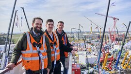 Drei Männer stehen auf einer Hebebühne über dem Freigelände der bauma.