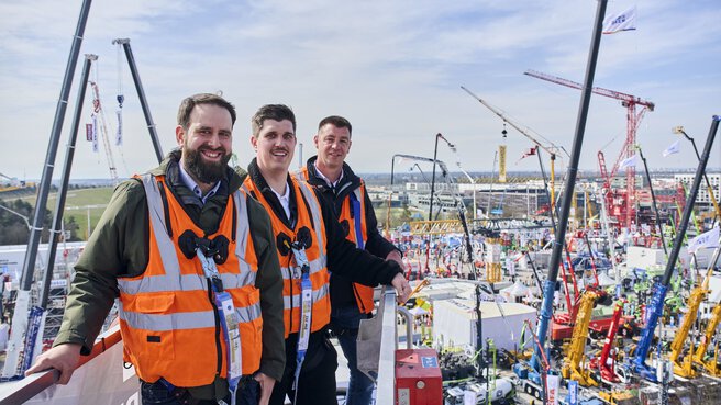Drei Männer stehen auf einer Hebebühne über dem Freigelände der bauma.