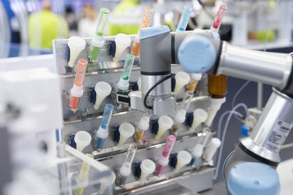 A laboratory robotic arm works with a series of test tubes organized in a rack. The test tubes contain different colored liquids. Laboratory equipment and people can be seen blurred in the background.
