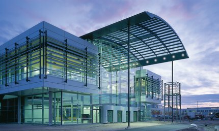 Exhibition entrance with glass and metal facade, large canopy and illuminated exterior facade at dusk and purple sky.