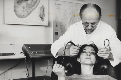 Ein altes schwarzweiß Foto von einem Arzt, der über eine Patientin gebeugt ist und medizinische Geräte an ihr Gesicht hält.