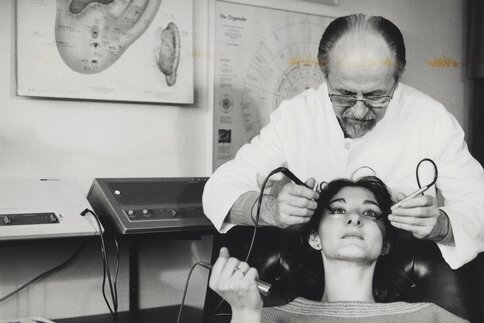 An old black and white photo of a doctor bent over a patient, holding medical equipment to her face.
