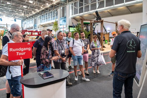 Eine Gruppe Personen hört während eines Rundgangs durch die Halle der INTERFORST einem Aussteller vor einer Präsentationstafel zu.