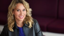 A smiling woman with shoulder-length blonde hair and blue eyes is wearing earrings and a necklace, a blue top and a black leather jacket.