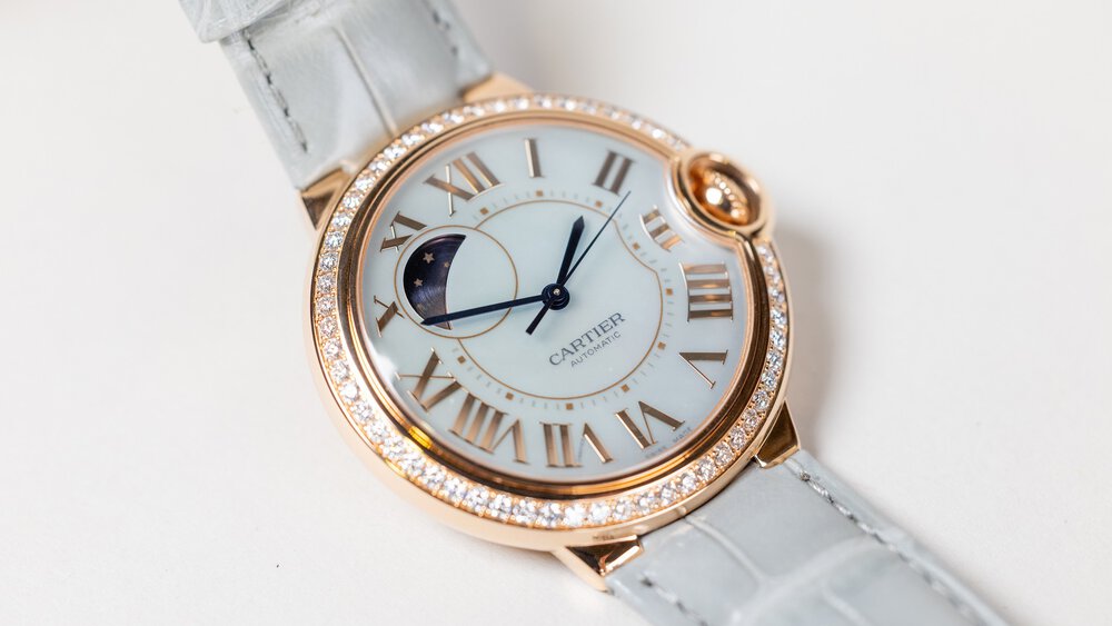 Close-up view of a delicate wristwatch with white straps, a gold-plated bezel set with gemstones, and a dial featuring the lunar cycle.