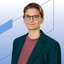 Eine lächelnde Frau mit kurzen braunen Haaren und Ohrringen trägt ein rötliches Oberteil und ein türkises Jackett darüber.