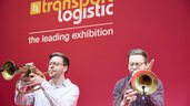 The two Brass Twins perform on horns on stage at the opening in front of a red transport logistic banner.