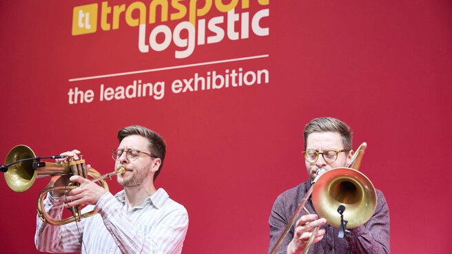 The two Brass Twins perform on horns on stage at the opening in front of a red transport logistic banner.
