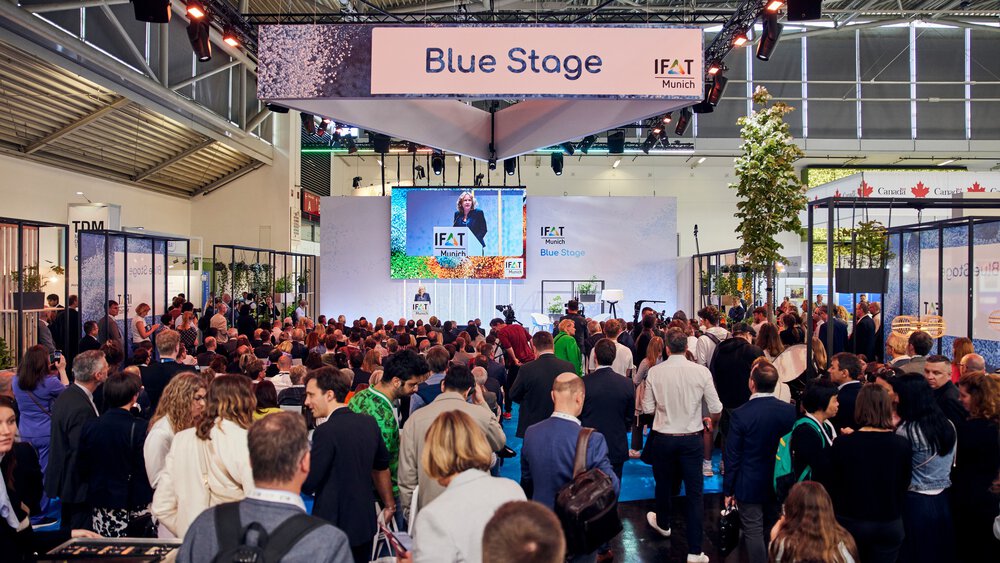 Large audience gathered in front of the “IFAT Munich – Blue Stage,” where a speaker stands at the podium; her talk is shown on a large screen above the stage, surrounded by exhibition booths and lighting elements.
