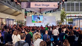 Large audience gathered in front of the “IFAT Munich – Blue Stage,” where a speaker stands at the podium; her talk is shown on a large screen above the stage, surrounded by exhibition booths and lighting elements.