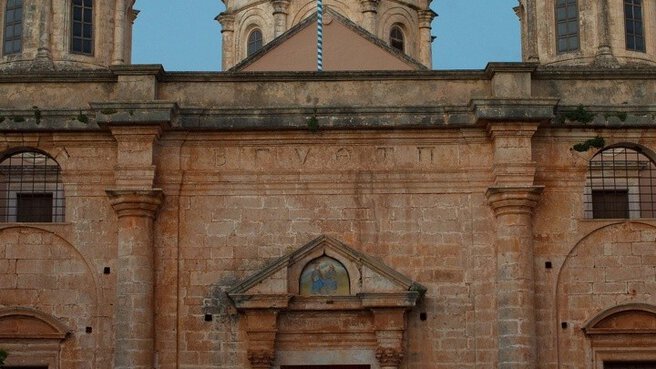 Mehrere Personen gehen bei Sonnenuntergang vor einer gewölbten Steinkirche, die von einem bogenförmigen Eingang eingerahmt wird.