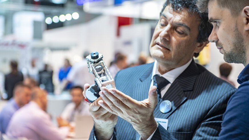 A man in a pinstripe suit presents a metallic device with several lenses to a trade fair visitor.