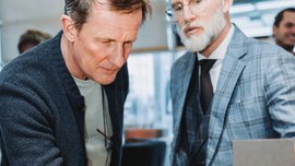 Two men with short hair, wearing suits and cardigans, are looking with interest at a watch displayed on a table.
