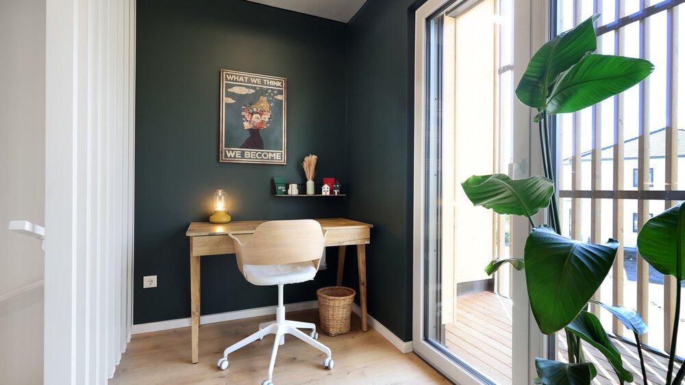 Ein kleines Heimbüro mit einem Holzschreibtisch, Stuhl, Lampe, Wandkunst, Zimmerpflanze und Zugang auf den Balkon.