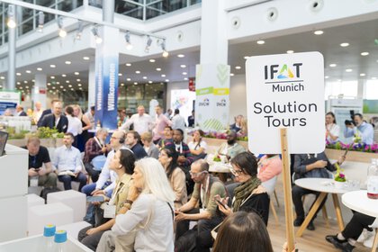 IFAT Munich Solution Tours sign held high, behind it a few people at tables and chairs can be seen out of focus.