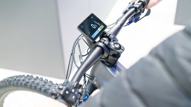 Nahaufnahme eines Fahrradlenkers mit moderner Digitalanzeige der von einer Person gehalten wird, vor neutralem Hintergrund.