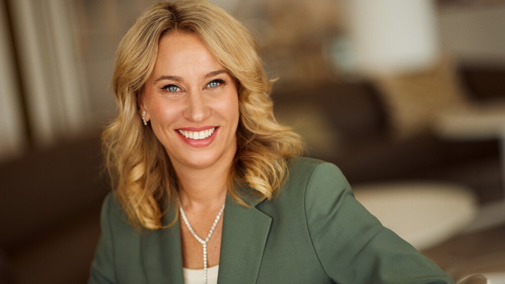 A smiling woman with long blonde hair and a white blouse under a green jacket. She is wearing a pearl necklace and earrings.