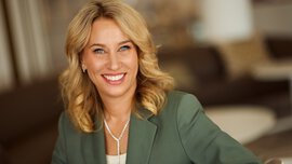 A smiling woman with long blonde hair and a white blouse under a green jacket. She is wearing a pearl necklace and earrings.
