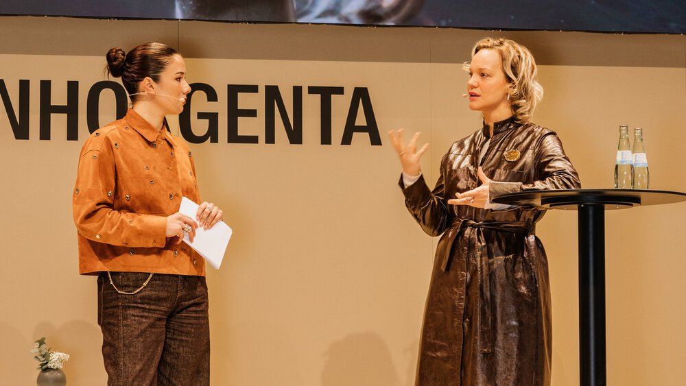 Two women are standing and talking on a stage, one is holding papers in her hand, behind them is a screen and a table with bottles of water.