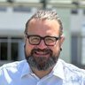 Ein lächelnder Mann mit Brille und Bart, der ein weißes Hemd trägt, steht draußen im Sonnenlicht.