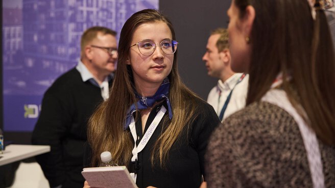 Eine Frau mit Brille und blauem Halstuch spricht auf der EXPO REAL mit einer anderen Teilnehmerin. Sie hält ein Notizbuch und eine Wasserflasche in der Hand und hört aufmerksam zu. Im Hintergrund sind weitere Personen in Gesprächssituationen zu sehen, die Messeatmosphäre wirkt konzentriert und professionell.