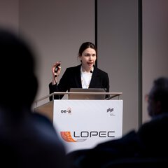 A woman in a black blazer is giving a speech from a podium bearing the inscription ‘LOPEC’ during a presentation; in the foreground, the audience is shown out of focus.