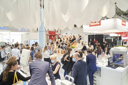 View of a well-filled exhibition stand with many people. In the background, some people are sitting on a kind of staircase and listening to a lecture.