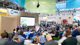 Trade visitors sitting in colorful armchairs at the BDE booth, following a presentation on a large screen.