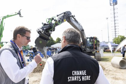 A man wearing a black vest with the words “CHANGE STARTS HERE” is being interviewed on the open-air site. Excavators are on display in the background.