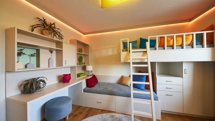 Stilvolles Schlafzimmer mit Hochbett, gemütlichen Sitzgelegenheiten, Schreibtisch und bunten Kissen. Warmes Licht und Pflanzen schmücken den Raum.