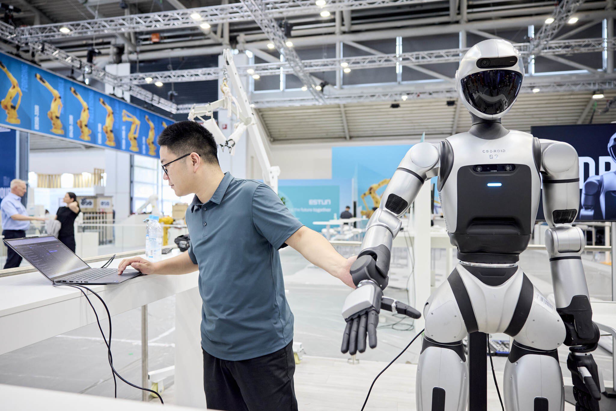 Ein Mann arbeitet an einem Laptop und hält dabei die Hand eines humanoiden Roboters. Die Szene spielt sich auf der Fachmesse automatica ab, im Hintergrund sind weitere Roboterarme und Messestände zu sehen.