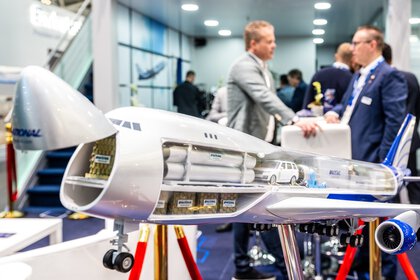 Model airplane with a cutout showing the cargo and car inside, displayed at a trade show with people chatting in the background.