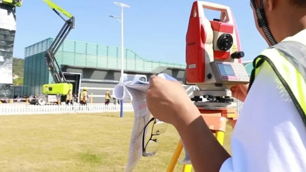 A man stands next to a surveying instrument in the open air in front of a construction site with a building crane and numerous workers running around.