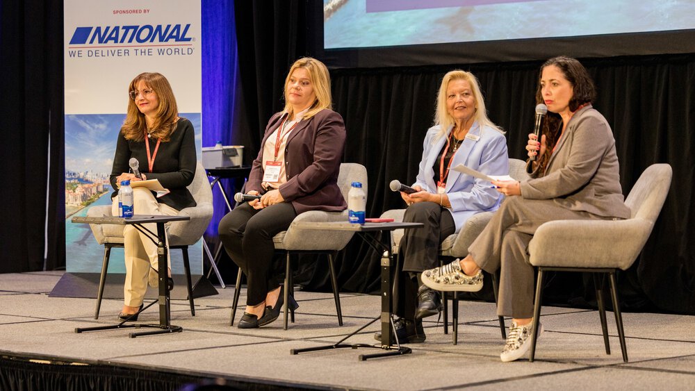 Four women with microphones sit on a stage and speak to an audience outside the frame. A sponsor's sign can be seen in the background.
