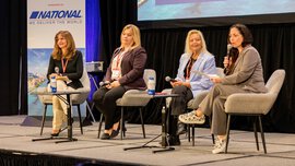Four women with microphones sit on a stage and speak to an audience outside the frame. A sponsor's sign can be seen in the background.