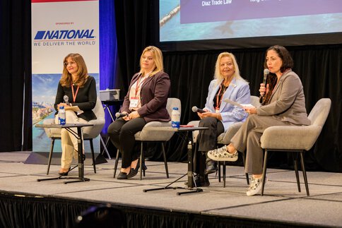 Four women with microphones sit on a stage and speak to an audience outside the frame. A sponsor's sign can be seen in the background.