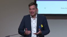 Ein Mann im dunkelblauen Blazer spricht am Podium, während hinter ihm ein Bildschirm mit einer Präsentationsfolie zeigt.