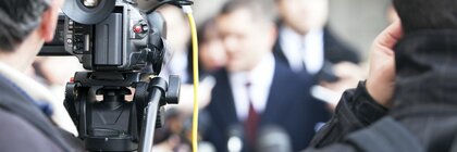 Eine Videokamera nimmt eine Gruppe von Menschen in Anzügen auf,  während einer Pressekonferenz oder eines Interviews.