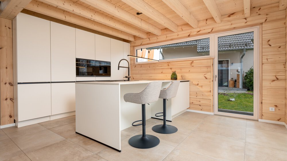 Moderne Küche mit Holzdecke und -wänden, cremefarbenen Schränken, Kochinsel mit Barhockern und großer Glastür zum Garten.